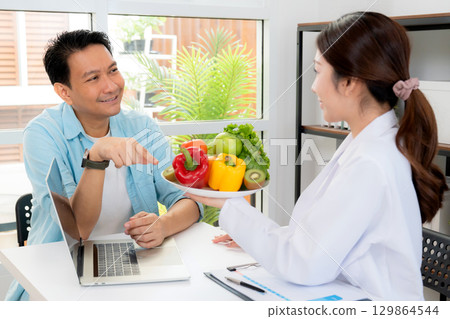 Asian nutritionist holding fresh fruits vegetables during consultation with patient in clinic. 129864544