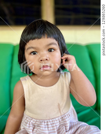 Portrait of a cute Asian little girl with big eyes sitting on the couch in living room 129865260