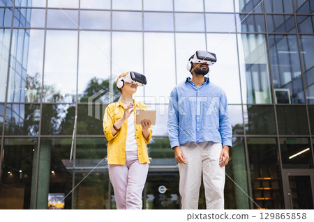 Two people wearing VR headsets explore augmented reality outdoors, near a modern glass building, with one holding a tablet. Two people wearing VR headsets explore augmented reality outdoors, near a modern glass building, with one holding a tablet. 129865858