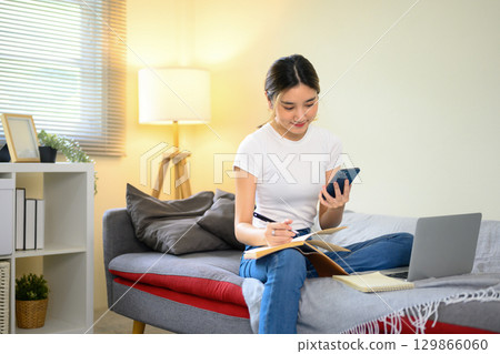 Young woman working from home, holding smartphone and writing notes in notebook 129866060