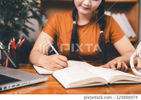 Asian girl doing homework and studying at home. woman using laptop at desk write notes watching webinar, study online, look at learn web class or having virtual call meeting remote working from home. 129866974