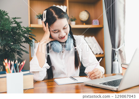Asian girl student wear headphones learning online watching webinar class looking at laptop computer eLearning remote lesson making notes or video calling virtual conference meeting teacher at home. Asian girl student wear headphones learning online watching webinar class looking at laptop computer eLearning remote lesson making notes or video calling virtual conference meeting teacher at home. 129867043