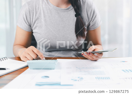 Young Asian woman making stack of coin. invest save finance concept, saving money, investment. dollar bills on table with pile of coins and banks calculator, money to save and invest to make income. 129867275
