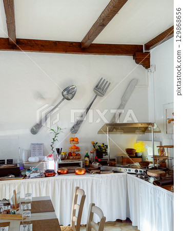 Restaurant interior features large wall decor of spoon, fork, and knife above a vibrant counter setup with fresh oranges and kitchen utensils, creating a unique dining atmosphere. Restaurant interior features large wall decor of spoon, fork, and knife above a vibrant counter setup with fresh oranges and kitchen utensils, creating a unique dining atmosphere. 129868656