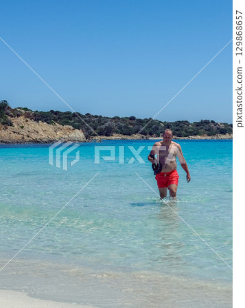 Man walking out of turquoise sea on a sunny day. Clear water, sandy beach, and rocky coastline in Sardinia. Concept of summer holidays, relaxation, seaside recreation, and healthy lifestyle Man walking out of turquoise sea on a sunny day. Clear water, sandy beach, and rocky coastline in Sardinia. Concept of summer holidays, relaxation, seaside recreation, and healthy lifestyle 129868657