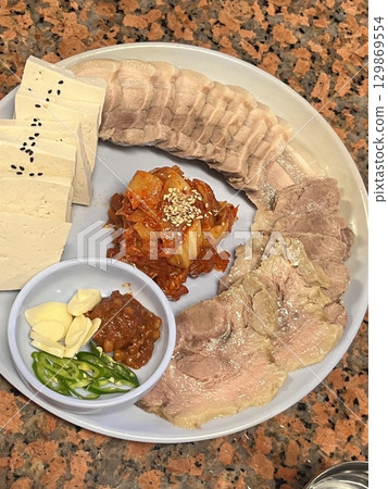 Tender slices of bossam, Korean boiled pork belly, served with fresh lettuce, garlic, and ssamjang. This flavorful and savory dish is perfect for wrapping and sharing in a traditional Korean meal. 129869554