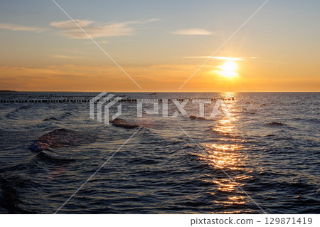 Scenic warm sea sunset view golden sunset over Baltic Sea reflects across rippling waves, creating a glowing path of light. Wooden breakwaters stretch into horizon calm rhythm to tranquil seascape Scenic warm sea sunset view golden sunset over Baltic Sea reflects across rippling waves, creating a glowing path of light. Wooden breakwaters stretch into horizon calm rhythm to tranquil seascape 129871419