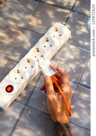 Person holding charger plug over white electricity extension cord with multiple sockets. Person holding charger plug over white electricity extension cord with multiple sockets. 129872820