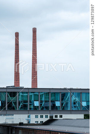 Industrial architecture featuring modern glass building with reflections, contrasting against iconic red smokestacks under a cloudy sky, Strijp S district in Eindhoven 129873897