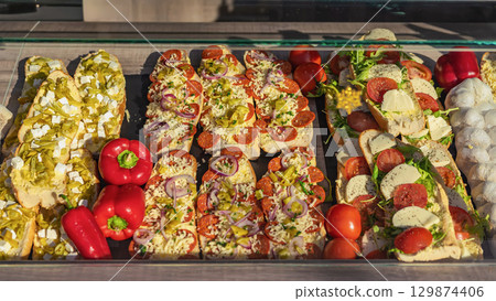 Baguette sandwiches for sale, fair street market stall. Assorted open faced baguettes topped with cheese, pepperoni, vegetables, feta on display. Concept of toppings, vegetarian option, street snack 129874406