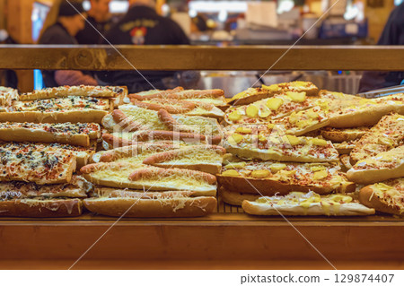 Baked baguettes filled with sausages, cheese, and toppings displayed at food stall. Concept of Christmas market, market food, holiday snacks, and street-style baking 129874407