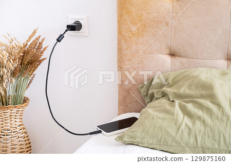 Phone charging cable connected to wall socket next to bed. 129875160