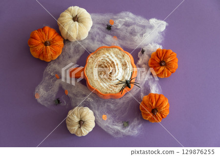 Halloween themed flat lay with a pumpkin latte, decorative pumpkins and spiderwebs on a purple background. 129876255