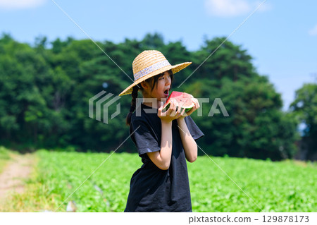 A scene of an elementary school girl wearing a straw hat holding a watermelon on a summer plateau 129878173