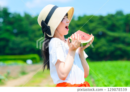 A scene of an elementary school girl wearing a straw hat holding a watermelon on a summer plateau 129878539