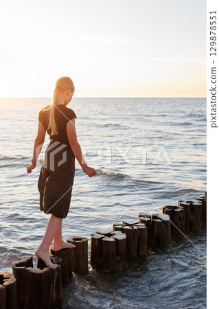 Cute adorable blond teenager happy kid girl walking by wooden breakwater posts at Baltic sea scenic warm summer sunset light. Balancing reflects challenges, choices and uncertainties of growing up 129878551