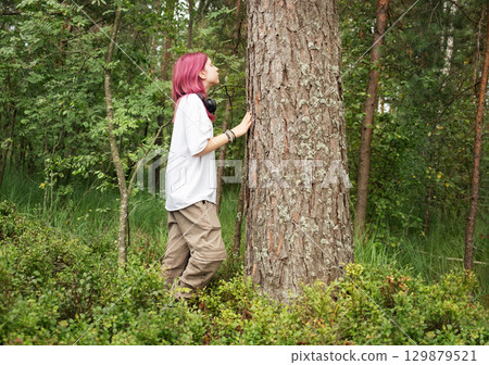 Young woman with pink hair embracing a tree in a forest, enjoying nature and tranquility 129879521