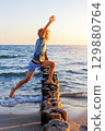 Boy leaps barefoot over wooden breakwater at seaside in golden evening light. Dynamic moment symbolizes childhood freedom, courage careless energetic leap toward unknown future 129880764