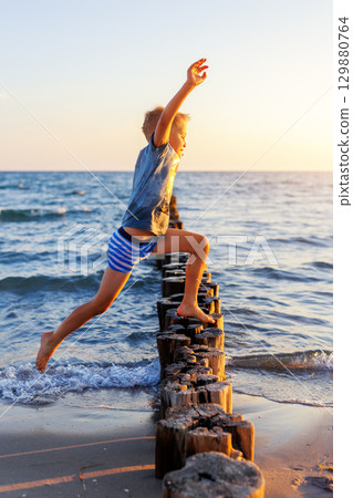Boy leaps barefoot over wooden breakwater at seaside in golden evening light. Dynamic moment symbolizes childhood freedom, courage careless energetic leap toward unknown future 129880764