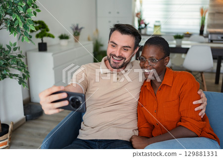 Happy multi-ethnic couple taking a selfie on the sofa at home 129881331