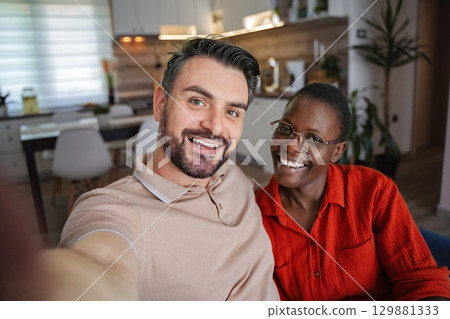 Happy multi-ethnic couple taking a selfie at home, smiling and looking at camera 129881333