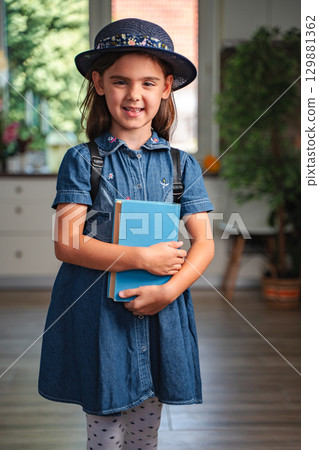 Smiling schoolgirl holding book and wearing backpack at home 129881362