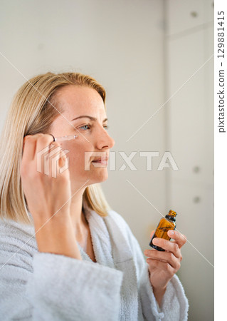 Woman applying essential oil on her face in bathroom 129881415