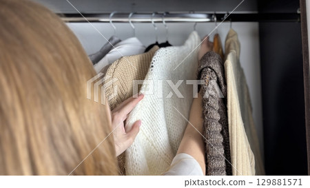 A woman sorts through clothes hanging on hangers in a closet. Reviewing and preparing seasonal wardrobe with warm knitted sweaters and jumpers for autumn winter. Minimalistic clothes organization 129881571