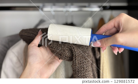 Woman using lint roller to clean lint laden clothes in closet, removing dust and debris from fabric. Wardrobe organization, clothing care, fashion and hygiene concept. Womens clothing maintenance 129881572