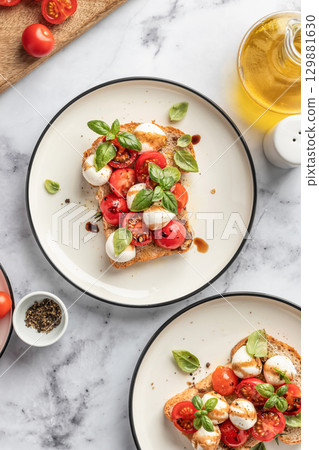 Caprese sandwich or toast with mozzarrella balls, cherry tomatoes, olive oil and fresh basil leaves top view. Healthy vegetarian breakfast, snack or lunch. White marble background 129881630