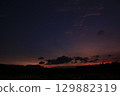 Peaceful rural scene of a rice field under a twilight sky. Dark clouds drift across a gradient of orange and purple hues, with silhouettes of trees and hills in the distance. 129882319