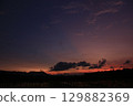 Wide view of a colorful sky transitioning from orange to purple above a flooded rice field at sunset. Dark clouds and distant mountain silhouettes create a peaceful mood. 129882369