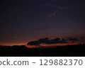 A moody twilight landscape featuring dark cloud shapes against a fading orange and purple sky. Silhouettes of distant hills and flooded rice fields evoke a calm rural scene. 129882370