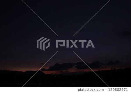 A quiet rural scene at nightfall with deep shadows over a rice field. The last light of sunset casts a subtle orange and purple hue behind dark clouds and silhouetted hills. 129882371