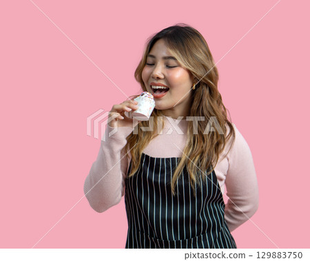 Young asian woman with an apron in celebratory or festive mood holding cupcake, decorated with white frosting and chocolate chip. Portrait on pink background with studio light, Isolated. Young asian woman with an apron in celebratory or festive mood holding cupcake, decorated with white frosting and chocolate chip. Portrait on pink background with studio light, Isolated. 129883750