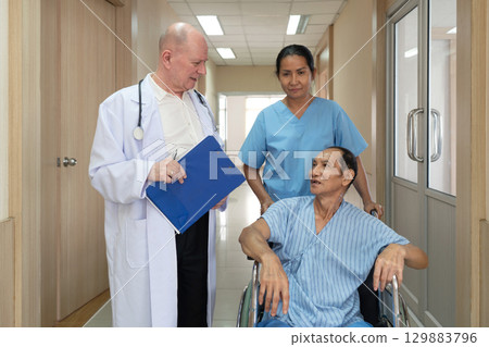 Medical staff engage with a patient in a wheelchair in a modern hospital corridor. A doctor and a nurse discuss the patient's condition attentively, ensuring clarity. 129883796