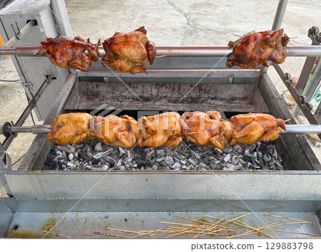 Juicy whole chicken are roasting on a rotisserie over charcoal at a street vendor stall. The smell of grilled meat fills the air, attracting customer in a lively market atmosphere. 129883798