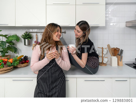 Two women with a striped apron drink coffee from mug while standing in front of counter in a modern kitchen. 129883801