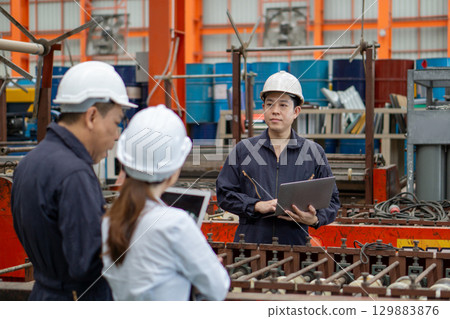 Three workers in a factory discuss a project using laptop computer. Everyone wearing protective hardhat. Atmosphere in manufactory with large machinery. Three workers in a factory discuss a project using laptop computer. Everyone wearing protective hardhat. Atmosphere in manufactory with large machinery. 129883876