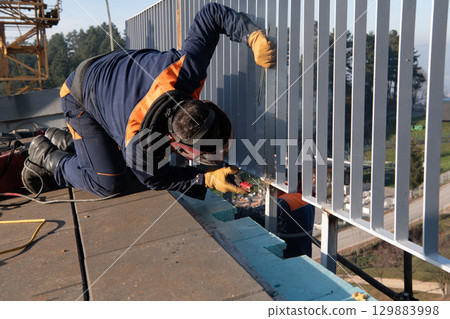 Industrial welder skillfully crafts a metal fence for a new business complex, using modern equipment that casts bright sparks and highlights the precision and dedication of the craft. 129883998