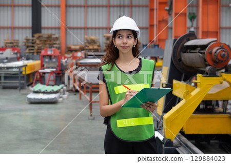 A woman wearing a safety vest and hardhat stand in a factory. She holds a clipboard and looks confident. A woman wearing a safety vest and hardhat stand in a factory. She holds a clipboard and looks confident. 129884023