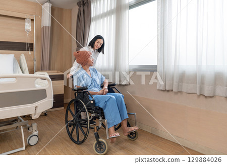 A caregiver support a chemo patient with knitted hat on her head sitting in wheelchair, they enjoy the natural light coming through the window in hospital room. The atmosphere is calm and comforting. 129884026