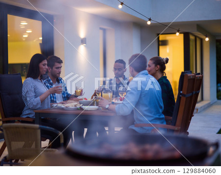 A group of young diverse people having dinner on the terrace of a modern house in the evening. Fun for friends and family. Celebration of holidays, weddings with barbecue 129884060