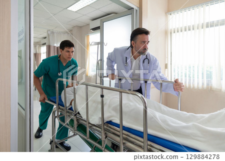 Two healthcare worker are hurriedly transferring a patient on a stretcher in a bright hospital emergency room during the day, illustrating urgency and teamwork in medical care. 129884278