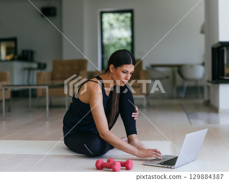 A young woman in sportswear is sitting in the living room and preparing for online training while using a laptop. 129884387