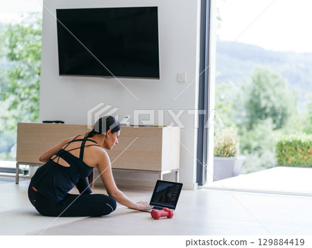 A young woman in sportswear is sitting in the living room and preparing for online training while using a laptop. A young woman in sportswear is sitting in the living room and preparing for online training while using a laptop. 129884419