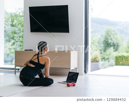 A young woman in sportswear is sitting in the living room and preparing for online training while using a laptop. A young woman in sportswear is sitting in the living room and preparing for online training while using a laptop. 129884432