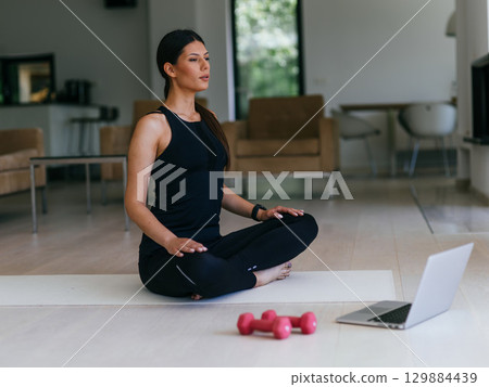 A young woman in sportswear is sitting in the living room and preparing for online training while using a laptop. A young woman in sportswear is sitting in the living room and preparing for online training while using a laptop. 129884439