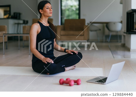 A young woman in sportswear is sitting in the living room and preparing for online training while using a laptop. 129884440