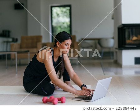A young woman in sportswear is sitting in the living room and preparing for online training while using a laptop. 129884442